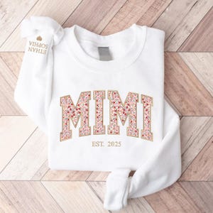 May include: White sweatshirt with the word "MIMI" in a floral design, outlined in gold. Below the word "MIMI" is the text "EST. 2025". The sleeve has the names "ETHAN SOPHIA" printed on it.