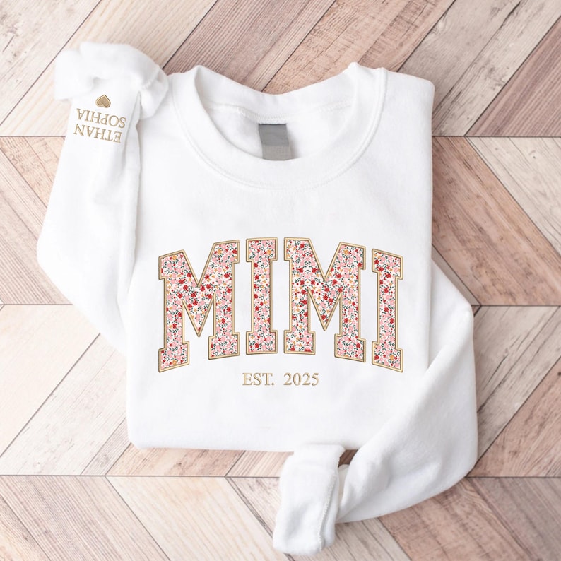 May include: White sweatshirt with the word "MIMI" in a floral design, outlined in gold. Below the word "MIMI" is the text "EST. 2025". The sleeve has the names "ETHAN SOPHIA" printed on it.