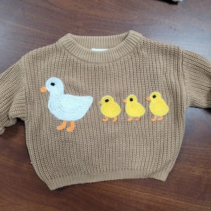 May include: A brown knit sweater with a crew neck and long sleeves. The sweater features an embroidered design of a white duck and three yellow chicks with orange feet. The sleeves are rolled up.