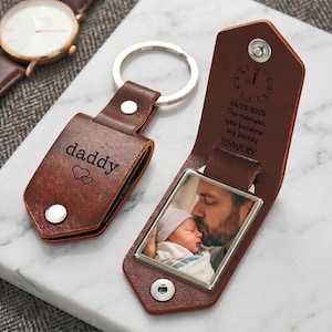 May include: A brown leather keychain with a metal ring and a snap closure. The exterior is engraved with the word daddy and two hearts. The interior features a photo of a person holding a baby, a clock face, the date 05/03/2026, and the text: The moment you became my Daddy, followed by a heart and the name AVERY.