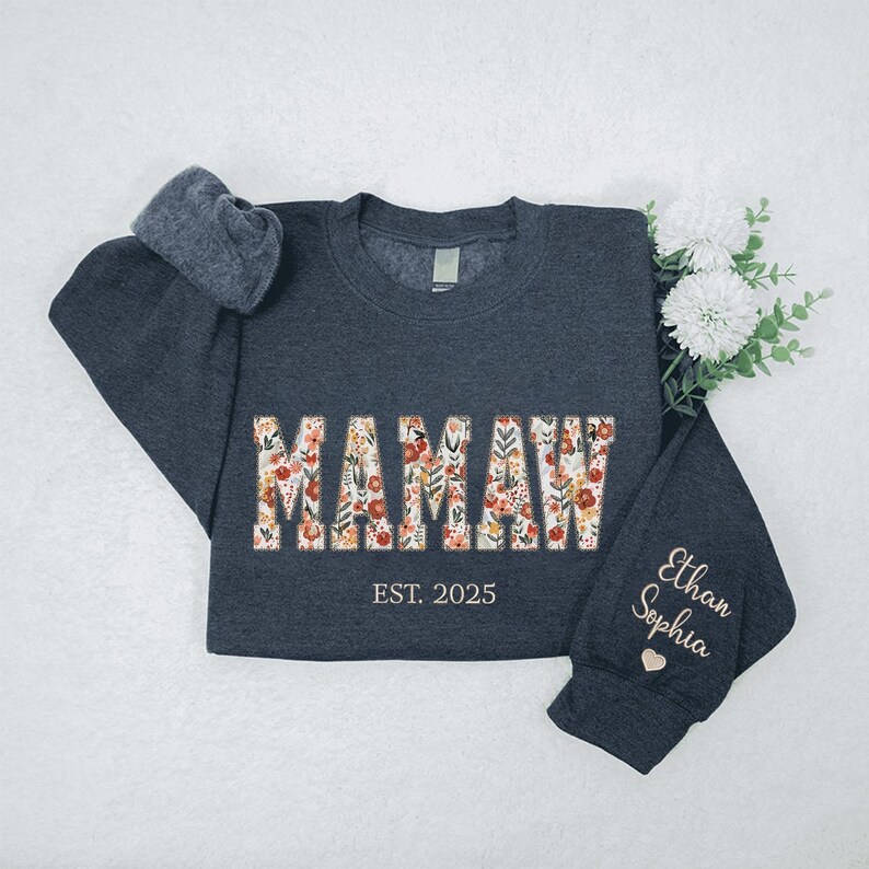 Op de afbeelding: Een donkergrijs sweatshirt met het woord "MAMAW" in bloemenprint. Het sweatshirt heeft de tekst "EST. 2025" onder het woord "MAMAW" en de namen "Ethan" en "Sophia" op de mouw.