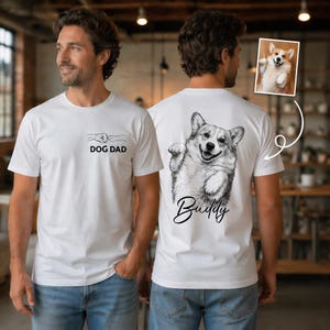 Pode incluir: Uma t-shirt branca com um gr&aacute;fico frontal de um punho e o texto DOG DAD. As costas exibem um grande esbo&ccedil;o a preto e branco de um Corgi com o nome Buddy escrito por baixo, correspondendo a uma pequena foto do mesmo c&atilde;o.