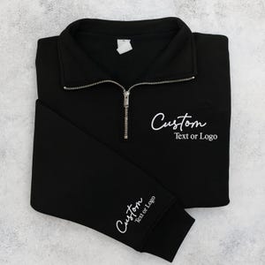 May include: Black quarter-zip sweatshirt with a silver zipper. The sweatshirt has the word "Custom" in white script on the chest and sleeve, with the text "Text or Logo" below. The sweatshirt is folded neatly on a light gray surface.