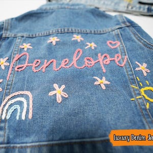 Puede incluir: Una chaqueta vaquera azul con detalles bordados, incluyendo un arco&iacute;ris, un sol, flores y el nombre "Penelope".