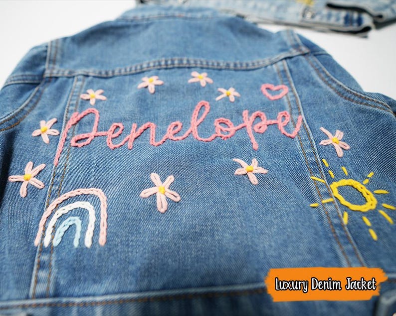 Puede incluir: Una chaqueta vaquera azul con detalles bordados, incluyendo un arco&iacute;ris, un sol, flores y el nombre "Penelope".