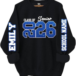 May include: Black sweatshirt with blue glitter text that reads "CLASS OF Senior 2026". The sleeves have the name "EMILY" and "SCHOOL NAME" in blue glitter. The sweatshirt has a crew neck and long sleeves.
