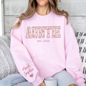May include: A light pink crewneck sweatshirt featuring the word AUNTIE in large block letters with a floral pattern. Below the word is the text EST. 2026. The sleeve displays the names Noah and Sophia in cursive script with a small pink heart.