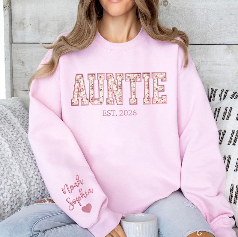 May include: A light pink crewneck sweatshirt featuring the word AUNTIE in large block letters with a floral pattern. Below the word is the text EST. 2026. The sleeve displays the names Noah and Sophia in cursive script with a small pink heart.