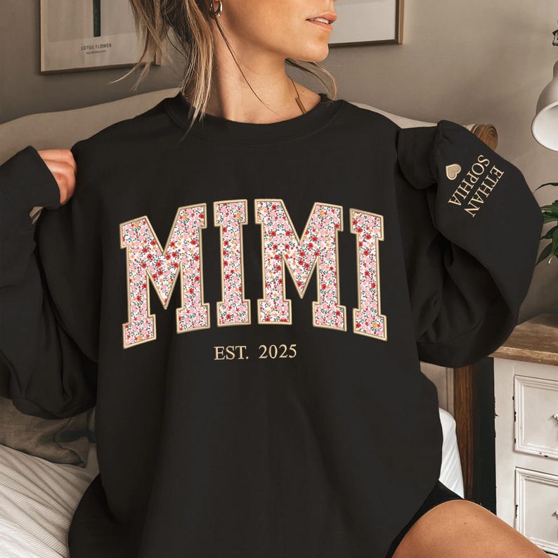 May include: Black sweatshirt with the word "MIMI" in a floral design, outlined in gold. The text "EST. 2025" is below the word. The sleeve has the names "ETHAN SOPHIA" embroidered. A cozy and personalized gift.