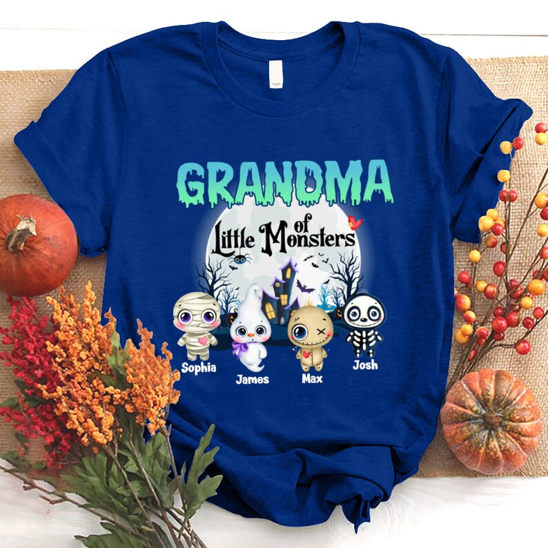 K&ouml;nnte beinhalten: Blaues T-Shirt mit dem Text "Grandma of Little Monsters" in Gr&uuml;n und Wei&szlig;. Das Shirt zeigt einen Mond mit einem Spukhaus und vier Cartoon-Monster: eine Mumie, ein Geist, eine Voodoo-Puppe und ein Skelett. Die Monster sind mit "Sophia", "James", "Max" und "Josh" beschriftet.