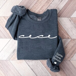 May include: A dark gray sweatshirt with the word "cece" written in white cursive on the front. The left sleeve has the names "Sophia Maria Jackson" written in white with a white heart below.