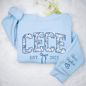 May include: Light blue sweatshirt with the name "CECE" in large, floral-patterned letters. The text "EST. 2025" and a bow graphic are below the name. The right sleeve has the names "Chris Sophia" and a heart symbol.