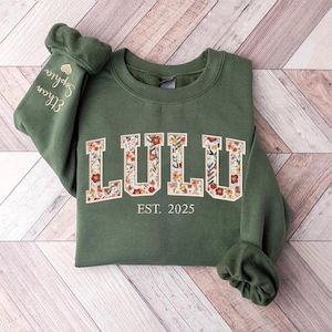 May include: A green sweatshirt with the word "LULU" in white letters outlined with a floral pattern. The text "EST. 2025" is below the word "LULU".