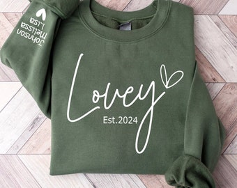 May include: An olive green sweatshirt with the word "Lovey" in a cursive font, a heart symbol, and "Est. 2024" printed in white. The left sleeve has the names "Melissa Lisa Johnson" and a heart symbol printed in white.