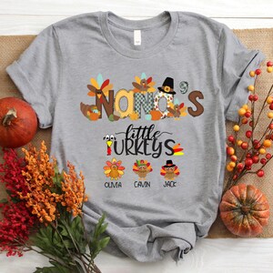 May include: Grey t-shirt with a Thanksgiving theme. The shirt features the text "Nana's Little Turkeys" with a turkey illustration for each child. The names Olivia, Gavin, and Jack are printed below the turkeys.
