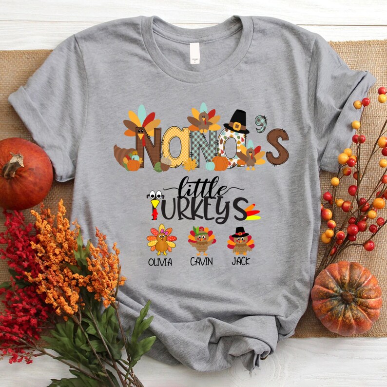 May include: Grey t-shirt with a Thanksgiving theme. The shirt features the text "Nana's Little Turkeys" with a turkey illustration for each child. The names Olivia, Gavin, and Jack are printed below the turkeys.