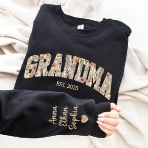 K&ouml;nnte beinhalten: Schwarzes Sweatshirt mit dem Wort "GRANDMA" in floralem Design und "EST. 2025" darunter. Der &Auml;rmel zeigt die Namen "Anna", "Ethan" und "Sophia" in Goldbuchstaben, mit einem kleinen Herzsymbol.