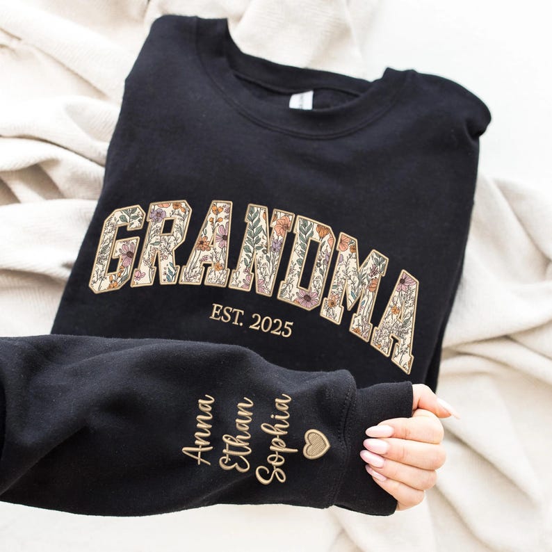 K&ouml;nnte beinhalten: Schwarzes Sweatshirt mit dem Wort "GRANDMA" in floralem Design und "EST. 2025" darunter. Der &Auml;rmel zeigt die Namen "Anna", "Ethan" und "Sophia" in Goldbuchstaben, mit einem kleinen Herzsymbol.
