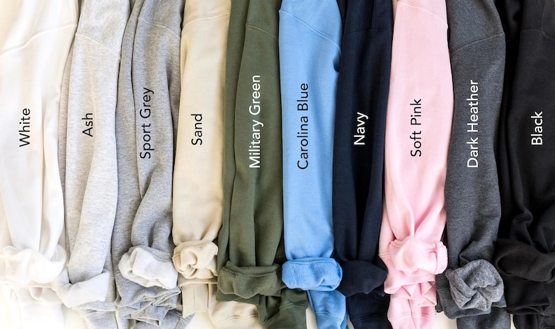May include: A row of ten folded sweatshirts in different colors, including white, ash, sport grey, sand, military green, carolina blue, navy, soft pink, dark heather, and black.