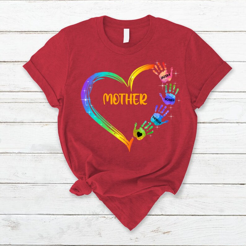 Pu&ograve; includere: Maglietta rossa con un cuore arcobaleno e impronte di mani. Il cuore contiene la parola "MAMMA". Le impronte di mani sono etichettate con i nomi Jaidira, Bobby e Teresa.