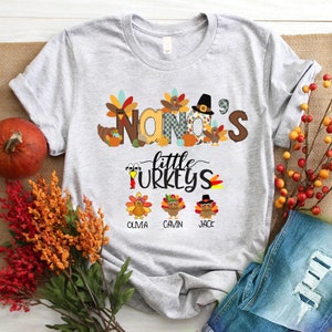 May include: A grey t-shirt with a Thanksgiving-themed design. The text "NANA'S little TURKEYS" is printed on the shirt, along with images of turkeys and autumn leaves. The names "OLIVIA", "CAVIN", and "JACK" are printed below the turkeys.