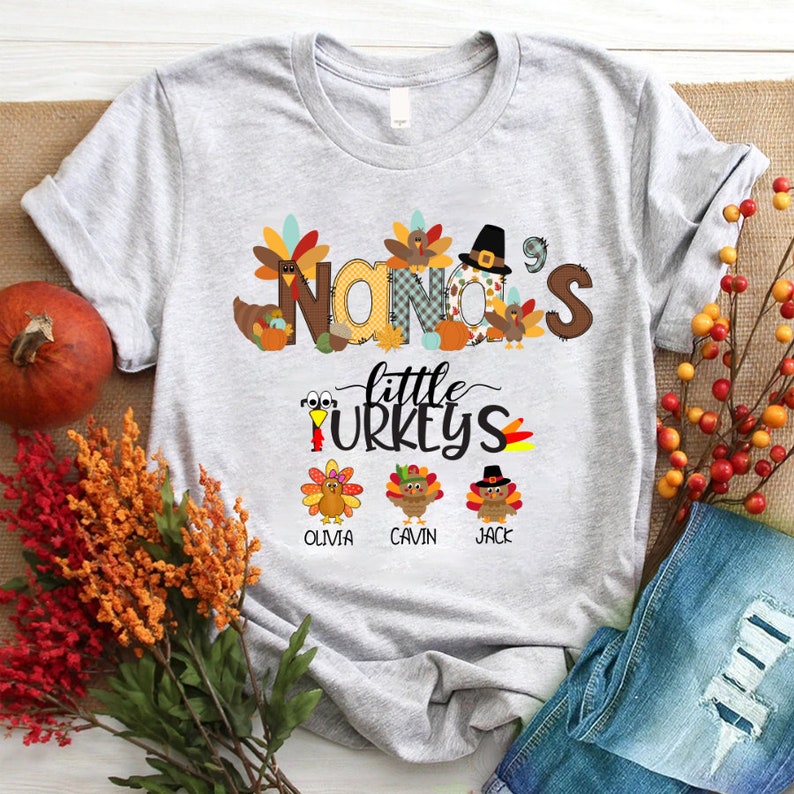 May include: A grey t-shirt with a Thanksgiving-themed design. The text "NANA'S little TURKEYS" is printed on the shirt, along with images of turkeys and autumn leaves. The names "OLIVIA", "CAVIN", and "JACK" are printed below the turkeys.