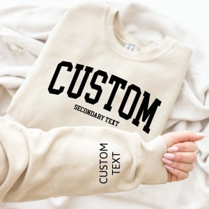 May include: A beige sweatshirt with black text that reads "CUSTOM" in a large font and "SECONDARY TEXT" in a smaller font. The sweatshirt also has the text "CUSTOM TEXT" on the sleeve.