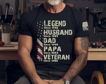 May include: A black t-shirt with a distressed American flag design and the text "LEGEND Since 1956", "HUSBAND Since 1989", "DAD Since 1990", "PAPA Since 2023", and "VETERAN Since 1986".