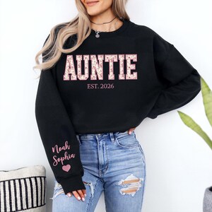 May include: A black long-sleeve sweatshirt featuring the word AUNTIE in large block letters with a pink floral pattern. Below the word is the text EST. 2026. The right sleeve displays the names Noah and Sophia in pink script above a pink heart.