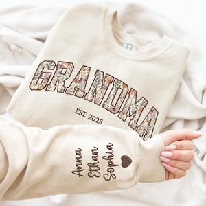 K&ouml;nnte beinhalten: Cremefarbenes Sweatshirt mit dem Wort "GRANDMA" in floralem Design. Darunter steht "EST. 2025". Auf dem &Auml;rmel sind die Namen "Anna", "Ethan" und "Sophia" sowie ein Herz gestickt. Ein personalisiertes Geschenk.