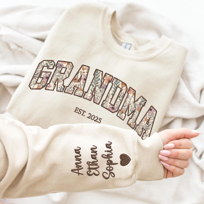 K&ouml;nnte beinhalten: Cremefarbenes Sweatshirt mit dem Wort "GRANDMA" in floralem Design. Darunter steht "EST. 2025". Auf dem &Auml;rmel sind die Namen "Anna", "Ethan" und "Sophia" sowie ein Herz gestickt. Ein personalisiertes Geschenk.