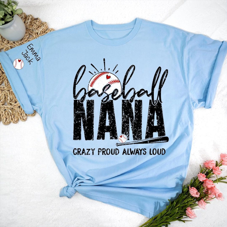 Peut inclure: T-shirt bleu clair sur le th&egrave;me du baseball avec les mots "Baseball Nana" en &eacute;criture noire et en lettres en bloc. Le t-shirt pr&eacute;sente un graphisme de baseball avec un c&oelig;ur et le texte "Crazy Proud Always Loud". La manche porte le nom "Emma Jack".