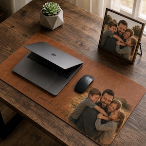 May include: A brown leather desk mat with a printed photo of a man and three children hugging. A laptop, mouse, and framed photo are on a wooden desk. The mat has the text "Our Favorite Guy."