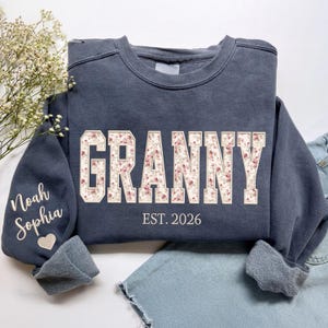 May include: A navy blue sweatshirt with the word "GRANNY" in a floral design across the chest. The text "EST. 2026" is below the word "GRANNY". The left sleeve has the names "Noah Sophia" and a heart.