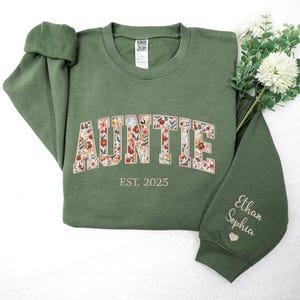 May include: A green sweatshirt with the word "AUNTIE" in floral letters. The text "EST. 2025" is below the word "AUNTIE". The right sleeve has the names "Ethan Sophia" embroidered on it.