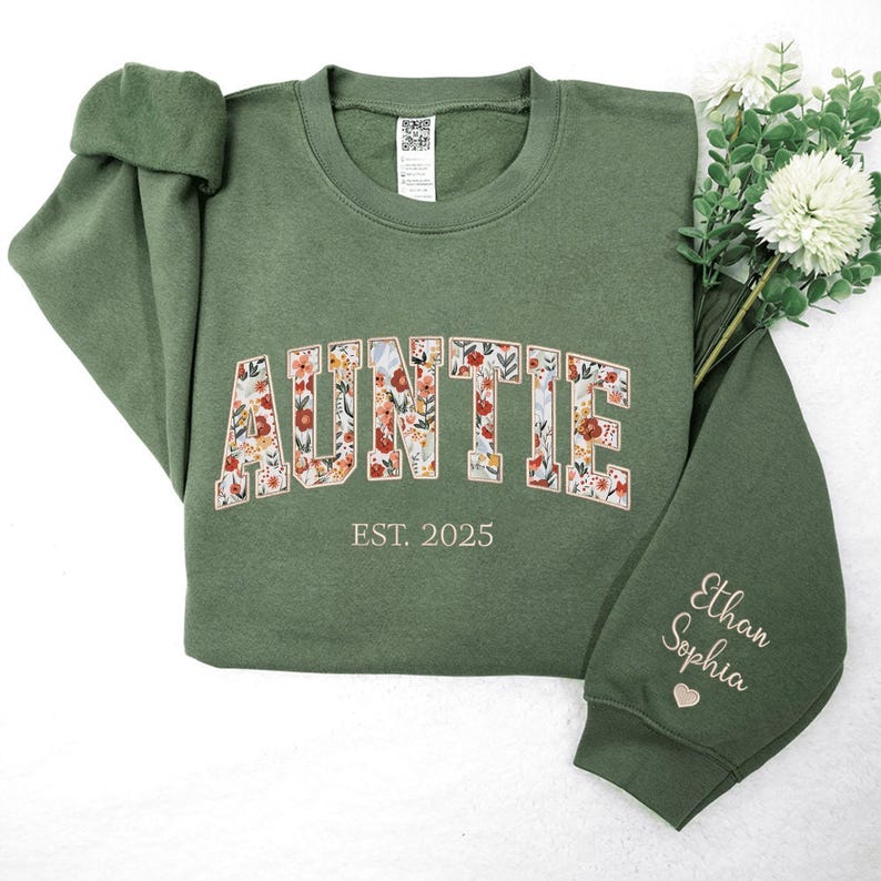 May include: A green sweatshirt with the word "AUNTIE" in floral letters. The text "EST. 2025" is below the word "AUNTIE". The right sleeve has the names "Ethan Sophia" embroidered on it.