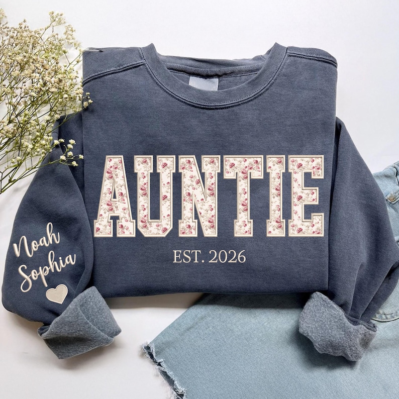 May include: A charcoal gray crewneck sweatshirt featuring the word AUNTIE in large block letters filled with a pink and cream floral pattern. Below the text is the phrase EST. 2026. The left sleeve displays the names Noah and Sophia with a heart.