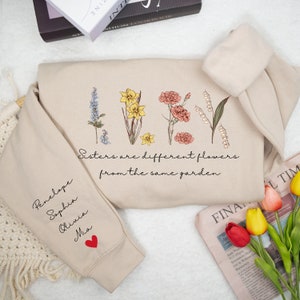 May include: A beige sweatshirt with a floral design and the text "Sisters are different flowers from the same garden". The sweatshirt has a red heart on the sleeve and the names Penelope, Sophia, Olivia, and Mia written on the sleeve.