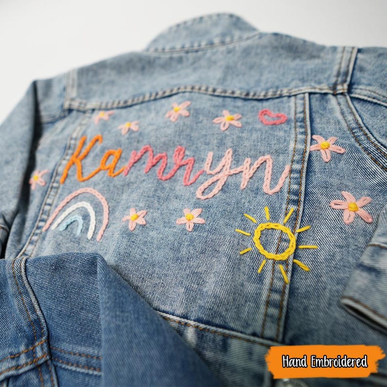 Puede incluir: Una chaqueta vaquera azul claro con el nombre "Kamryn" bordado en la espalda con hilo rosa, naranja y amarillo. El bordado tambi&eacute;n incluye un arco&iacute;ris, un sol y flores rosas.