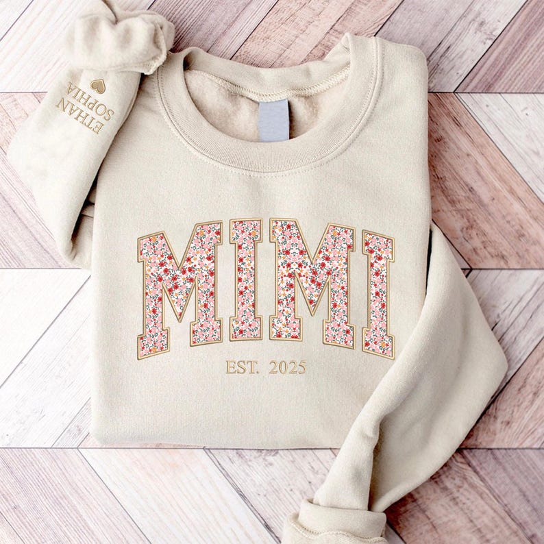 May include: A beige sweatshirt with the word "MIMI" in a floral design, with "EST. 2025" below. The sleeve cuff displays the names "ETHAN SOPHIA". The sweatshirt is folded on a wooden surface.