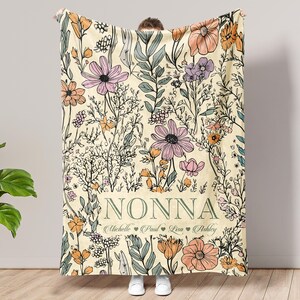 May include: A cream-colored blanket with a floral design featuring pink, orange, and purple flowers, and green leaves. The word "NONNA" is displayed in the center, with names below. The blanket is held up by a person.