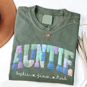 May include: Olive green t-shirt with the word "AUNTIE" in patchwork letters. The names "Sophia", "James", and "Noah" are written below. A gold necklace and a brown hat are also visible.