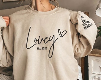May include: A beige sweatshirt with the text "Lovey Est. 2023" and a heart on the front. The sleeve has the names "Stephanie, Melissa, Lisa" with a heart at the end.