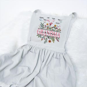 May include: A white apron with a floral design and the text "Grandma, we love you". The apron has two pockets and is decorated with butterflies and names.