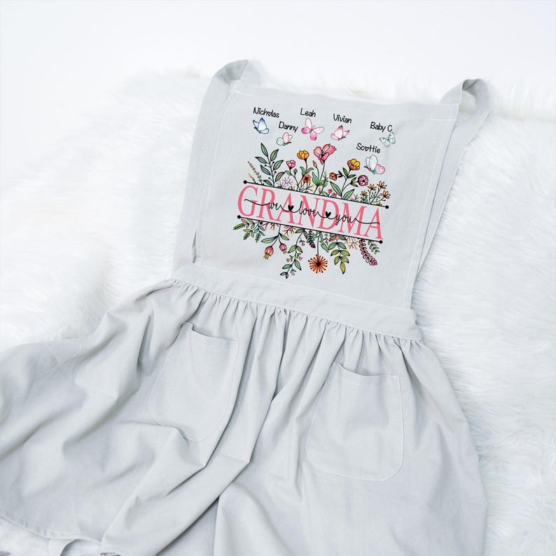 May include: A white apron with a floral design and the text "Grandma, we love you". The apron has two pockets and is decorated with butterflies and names.