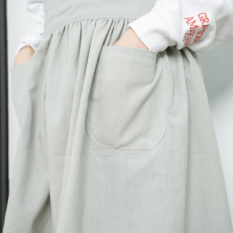May include: A light green pinafore dress with two large pockets on the front. The dress has a gathered skirt and a simple design.