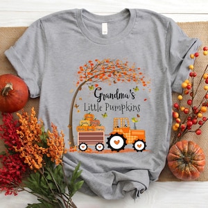 May include: Gray t-shirt with a fall-themed design. A tractor pulls a wagon full of pumpkins with names written on them. The text reads "Grandma's Little Pumpkins".
