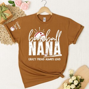 Peut inclure: Un t-shirt marron avec l'inscription blanche "baseball NANA" et un motif de baseball. Le texte "CRAZY PROUD ALWAYS LOUD" est en dessous. La manche porte l'inscription "Emma Jack" et un motif de baseball.