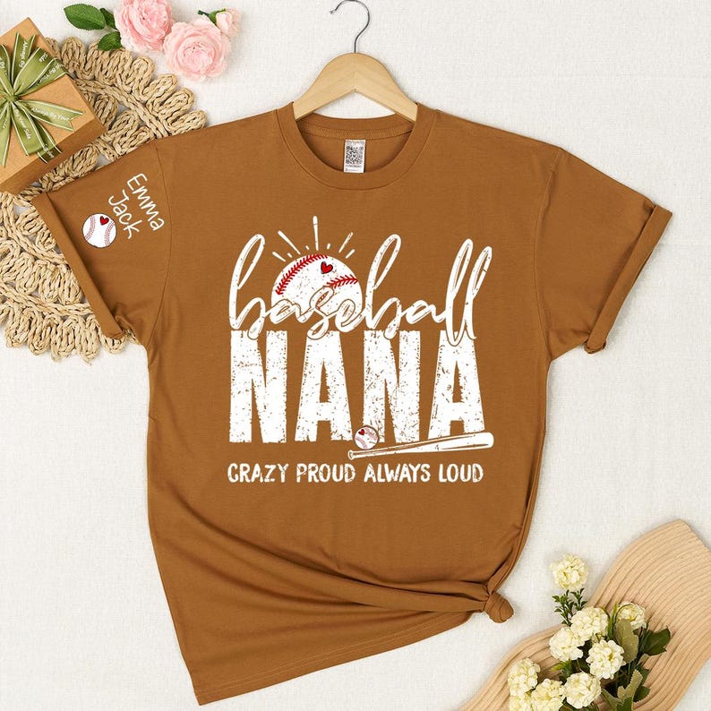 Peut inclure: Un t-shirt marron avec l'inscription blanche "baseball NANA" et un motif de baseball. Le texte "CRAZY PROUD ALWAYS LOUD" est en dessous. La manche porte l'inscription "Emma Jack" et un motif de baseball.