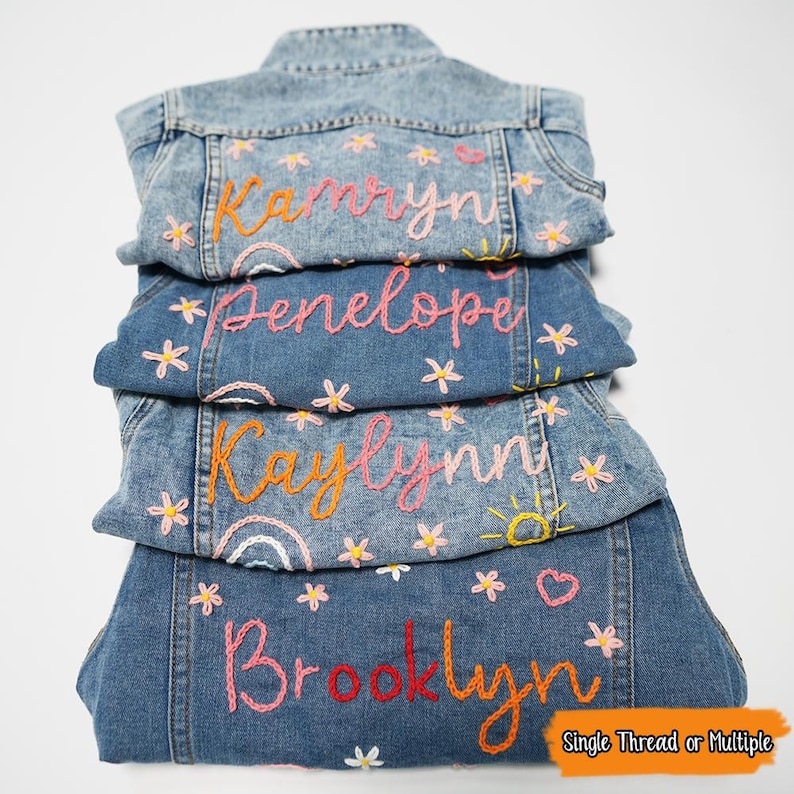 Puede incluir: Cuatro chaquetas vaqueras con nombres bordados en la espalda. Los nombres son "Kamryn", "Penelope", "Kaylynn" y "Brooklyn". Cada nombre est&aacute; bordado en diferentes colores con un arco&iacute;ris, un sol y flores.